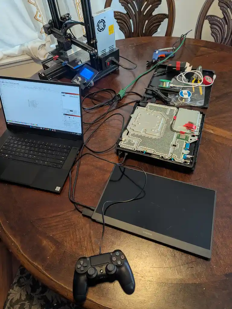 A cluttered wooden table holds a laptop displaying a design program, a disassembled gaming console with exposed internals, a…
