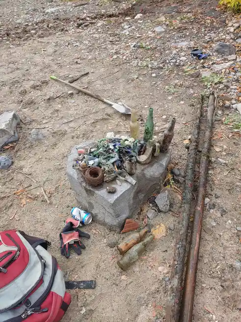 An outdoor scene shows a collection of old, dirty glass bottles and bottle fragments piled on a large grey stone. Nearby lie…
