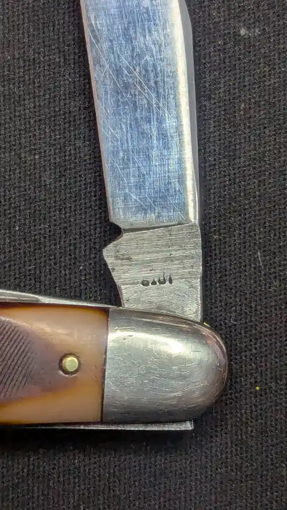 A close-up photo of a well-worn pocketknife blade resting on a dark textured surface, showing visible scratches and a small…