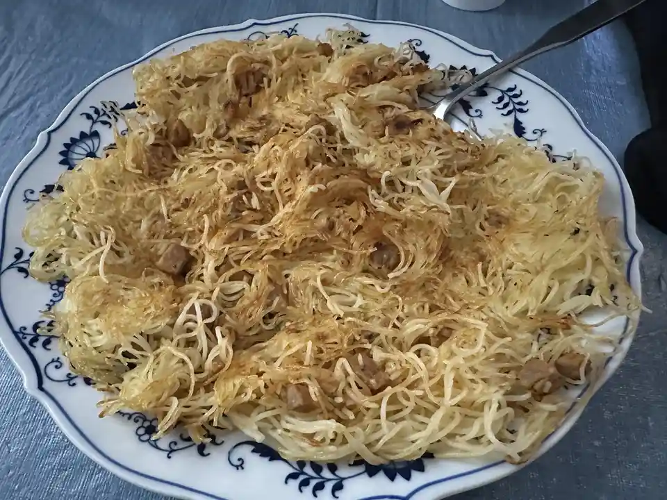 A close-up photo shows a plate of fried noodles with small pieces of meat, served on a white plate with blue floral…