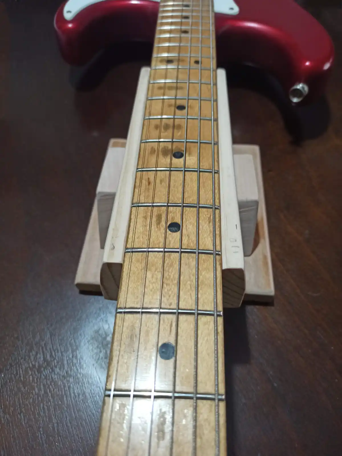 A close-up photo shows the fretboard of a red electric guitar resting on a homemade wooden stand. The guitar's neck is…