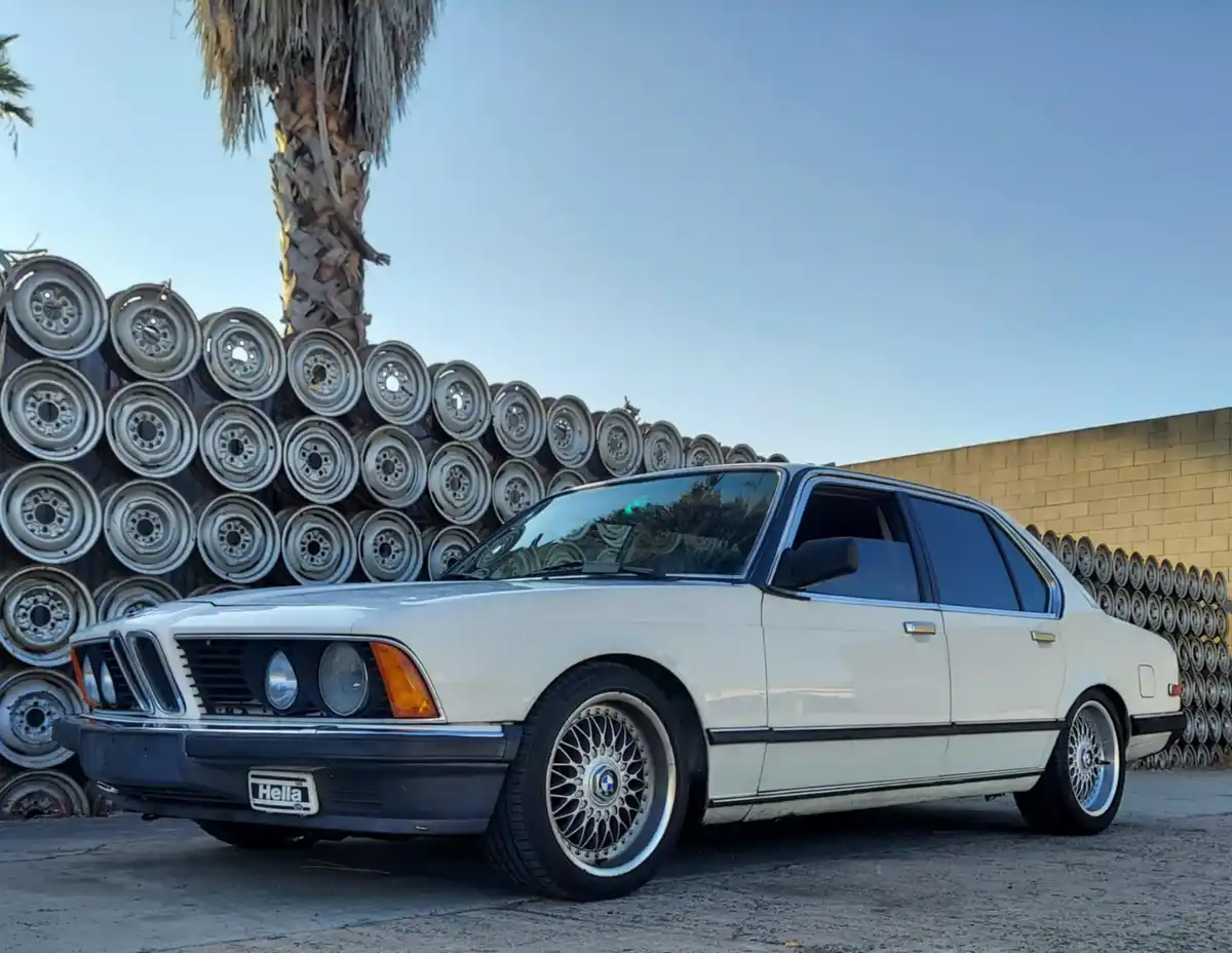 A white BMW E21 sedan is parked in front of a large stack of spare wheels, with a palm tree and a brick wall in the…