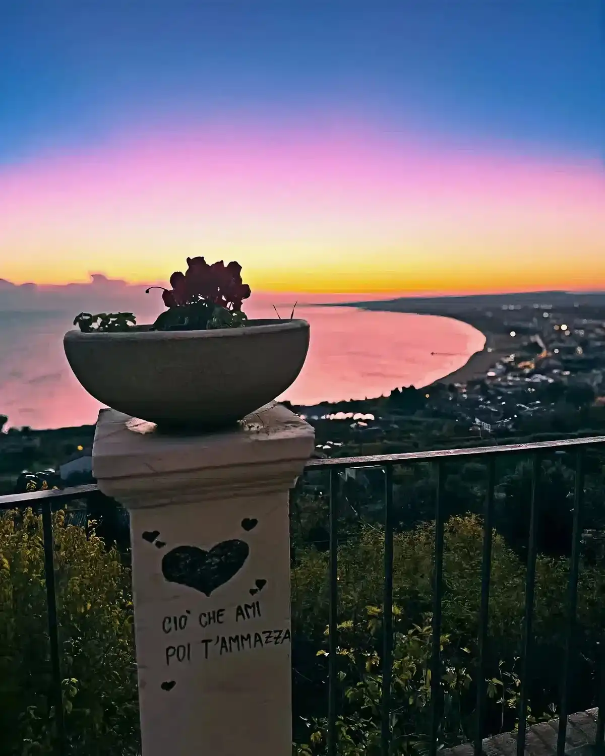 A serene sunset photo shows a stone pillar with a potted plant on top, overlooking a coastal town and bay. The pillar has a…