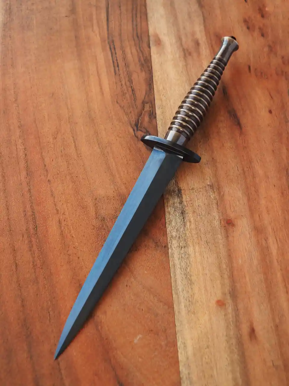 A close-up photo shows a dagger with a dark blue blade and a ribbed, metallic handle resting diagonally on a wooden surface…