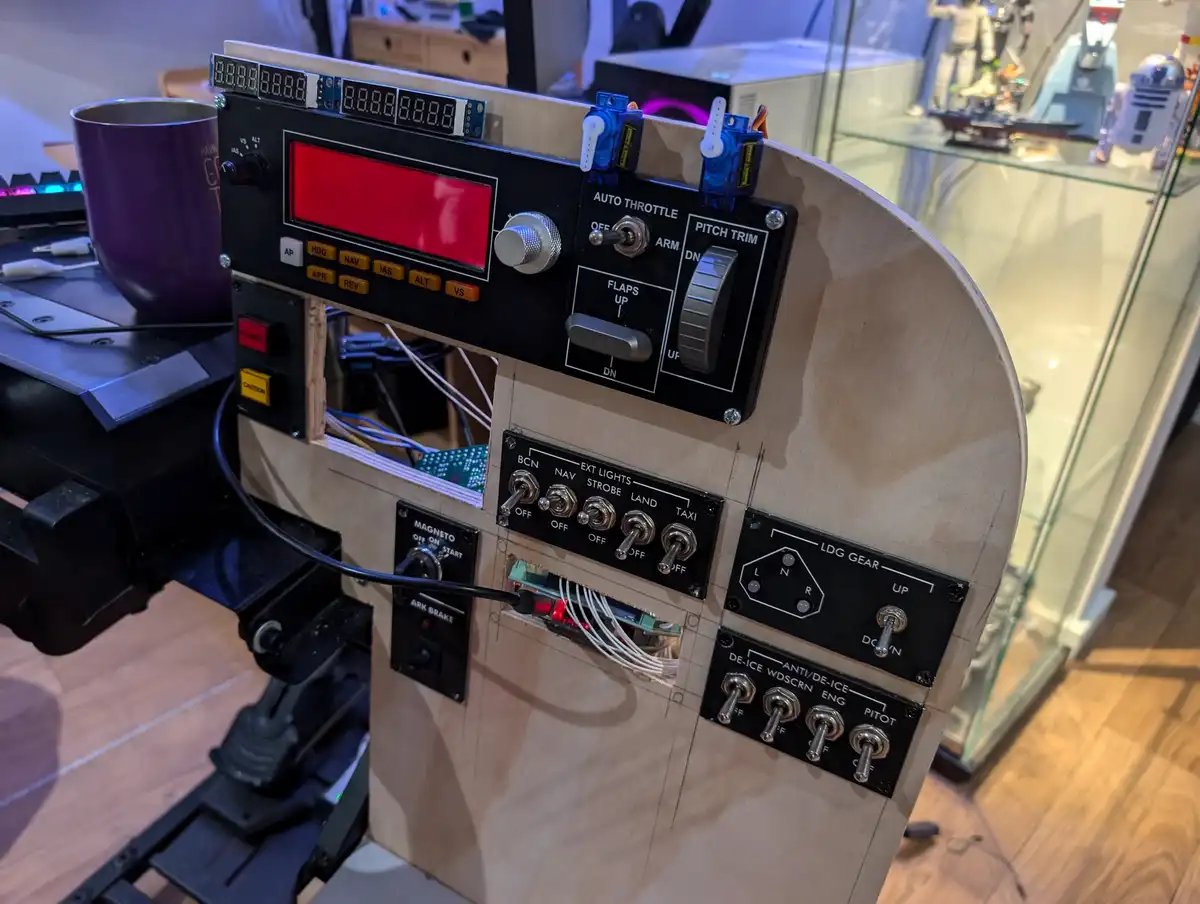 A homemade flight simulator cockpit with a red digital display, various switches, and knobs labeled with aviation terms like…
