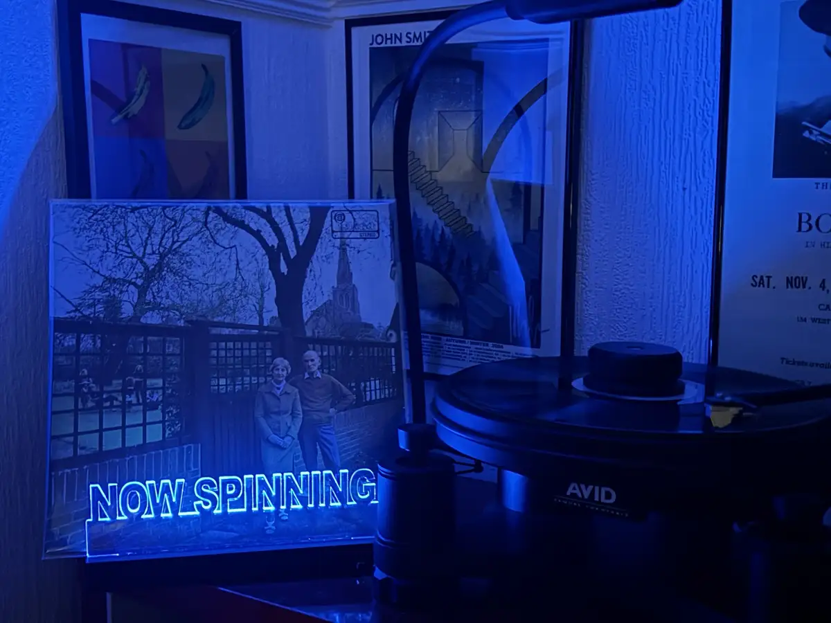 A vinyl record player with a glowing blue 'NOW SPINNING' sign is shown in a dimly lit room bathed in blue light. The record…