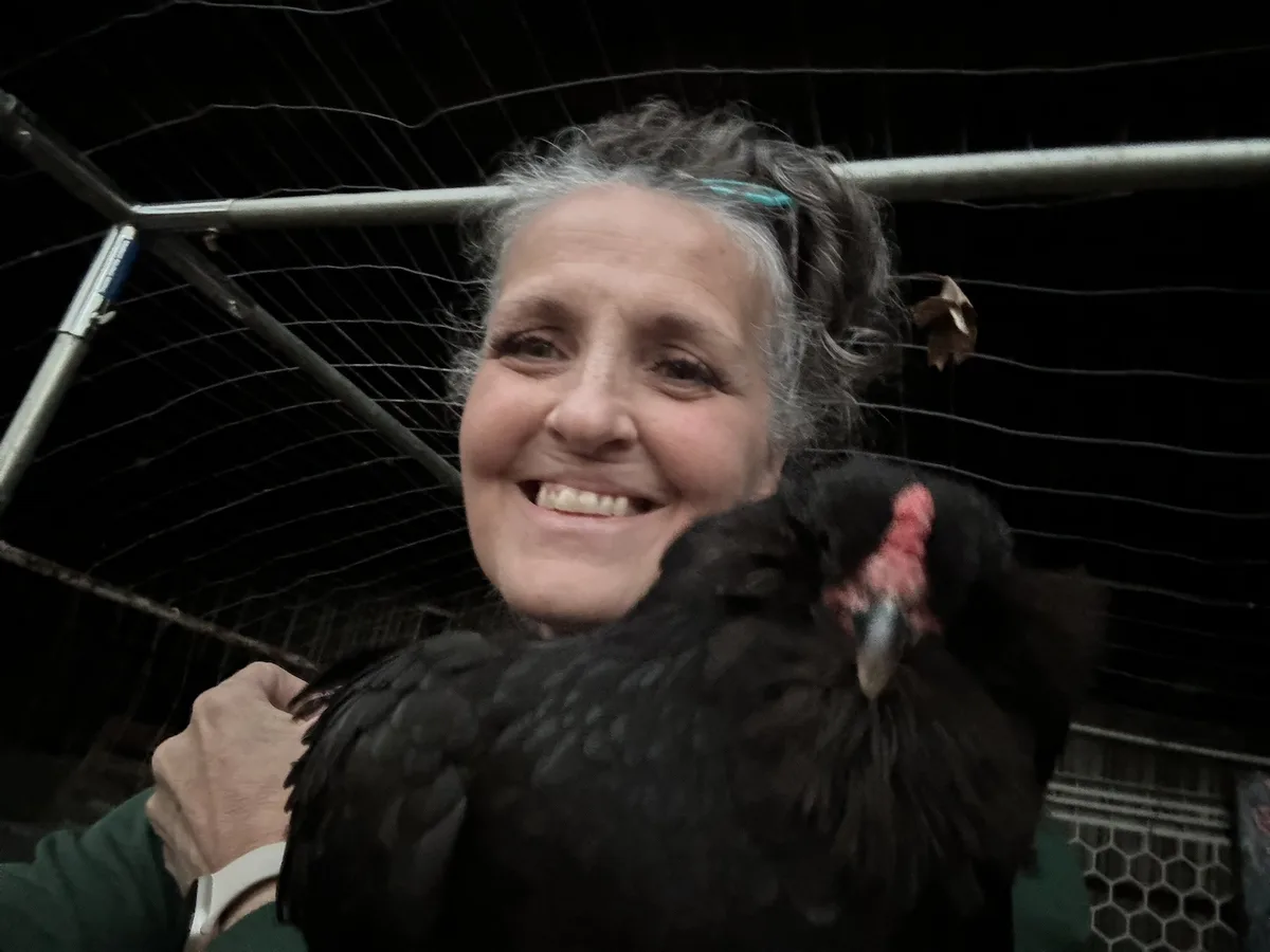 A smiling woman with gray hair holds a black chicken close to her face, partially obscuring it, in front of a wire mesh…