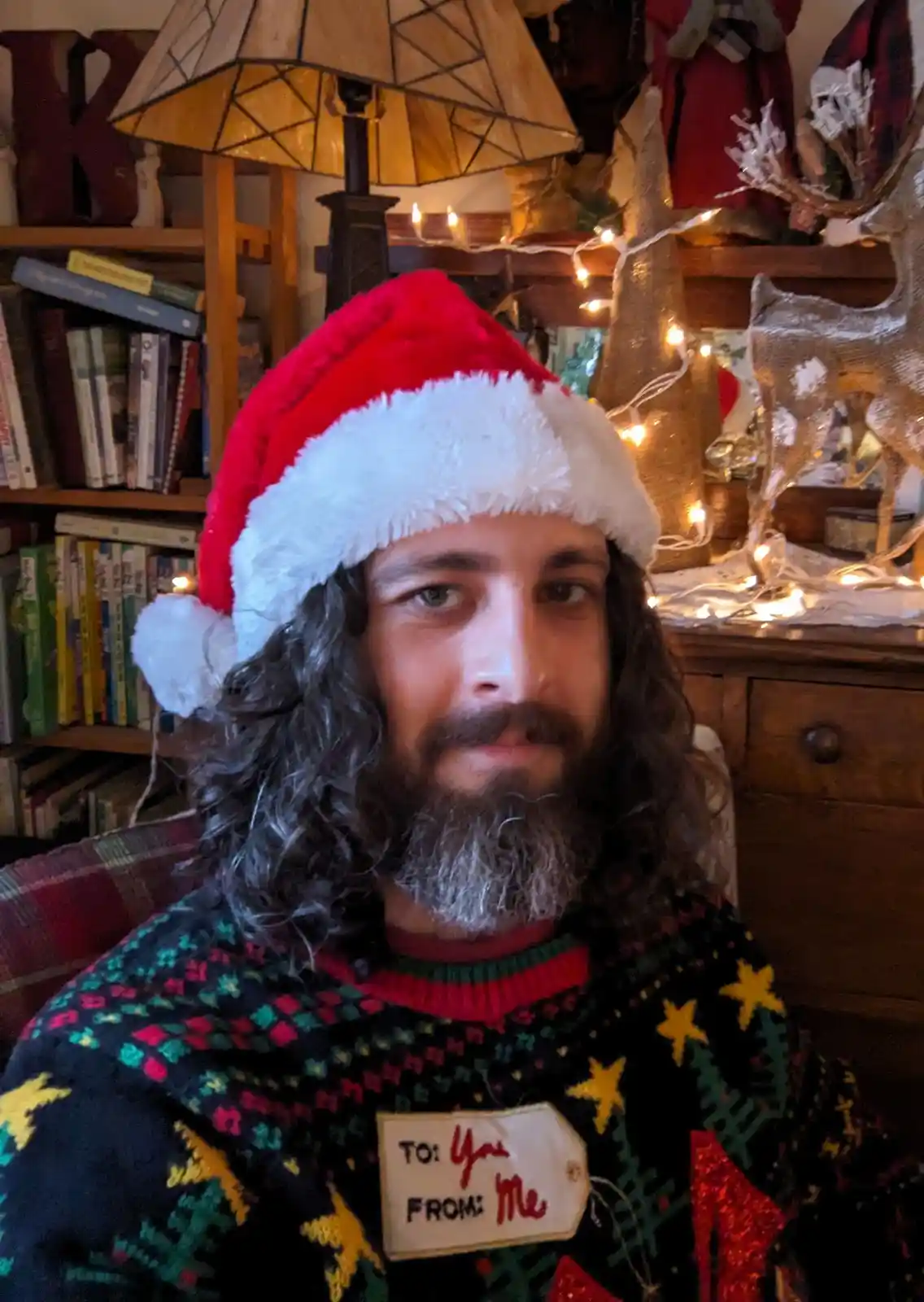 A man with long dark hair and a beard wears a red and white Santa hat and a colorful Christmas sweater with a tag that reads…