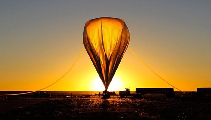 A hot air balloon is shown at sunrise or sunset, its envelope glowing orange against a gradient sky. The balloon is tethered…