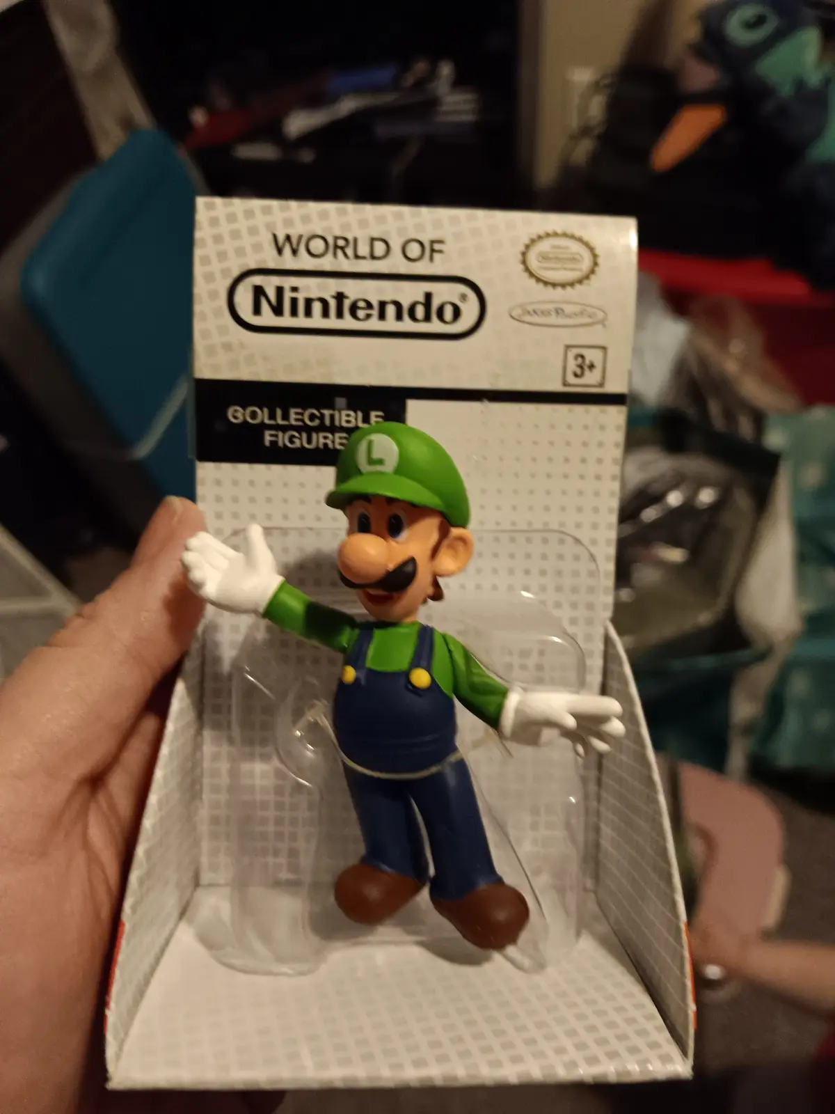 A hand holds a 'World of Nintendo' collectible figure box containing a Luigi action figure in his classic green hat and blue…