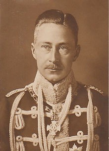 A sepia-toned vintage portrait of a man in formal military uniform with medals and epaulets, looking directly at the camera…