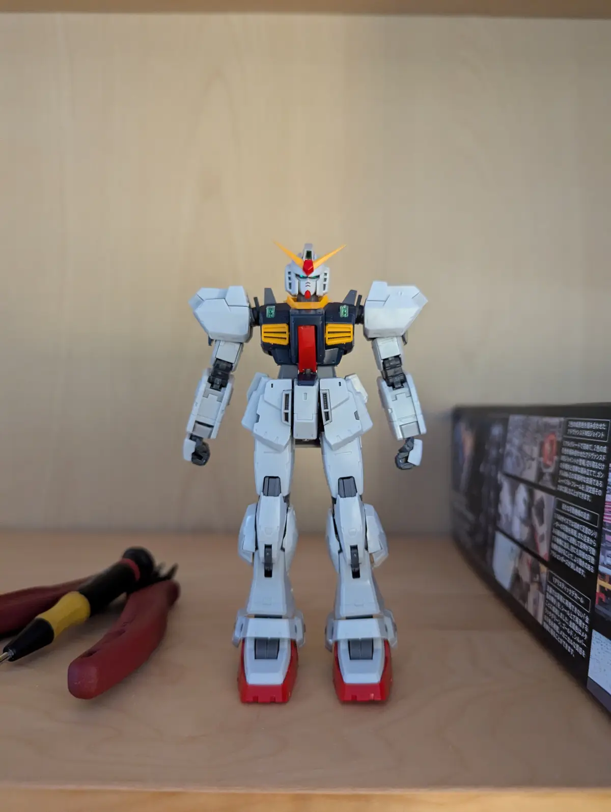 A white, red, and yellow plastic robot model stands upright on a wooden shelf, with a pair of red-handled pliers and a…