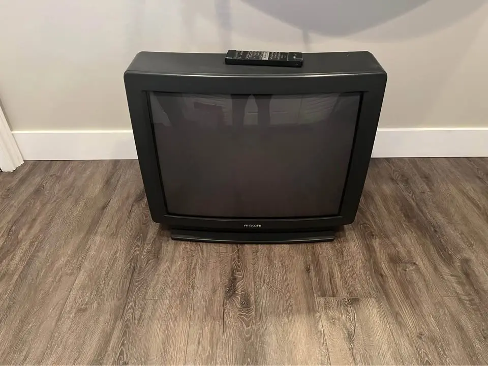 A black Hitachi CRT television sits upright on a wood-patterned floor with a remote control resting on top. The screen is…
