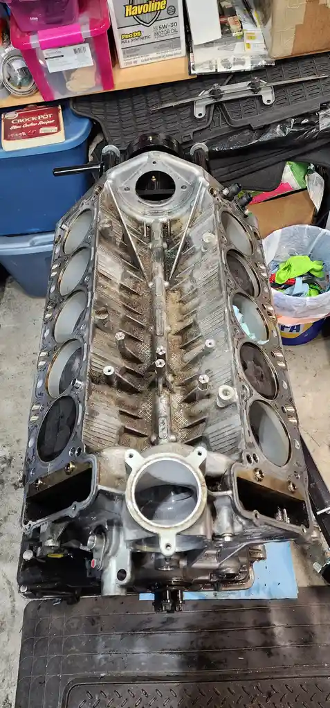 A dirty, disassembled V8 engine block sits on a workbench in a cluttered garage, surrounded by tools, oil containers, and…