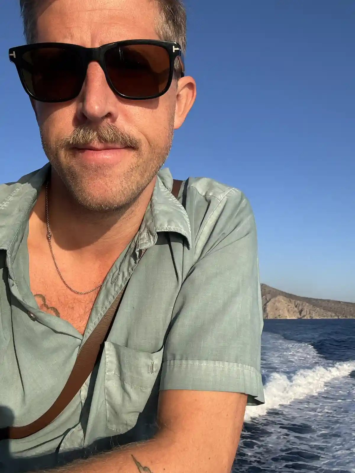 A man with a mustache and sunglasses, wearing a light green shirt and a necklace, takes a selfie on a boat with the ocean…