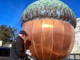 A person wearing a black cap and jacket is standing next to a giant, copper-colored acorn sculpture with a green, textured…