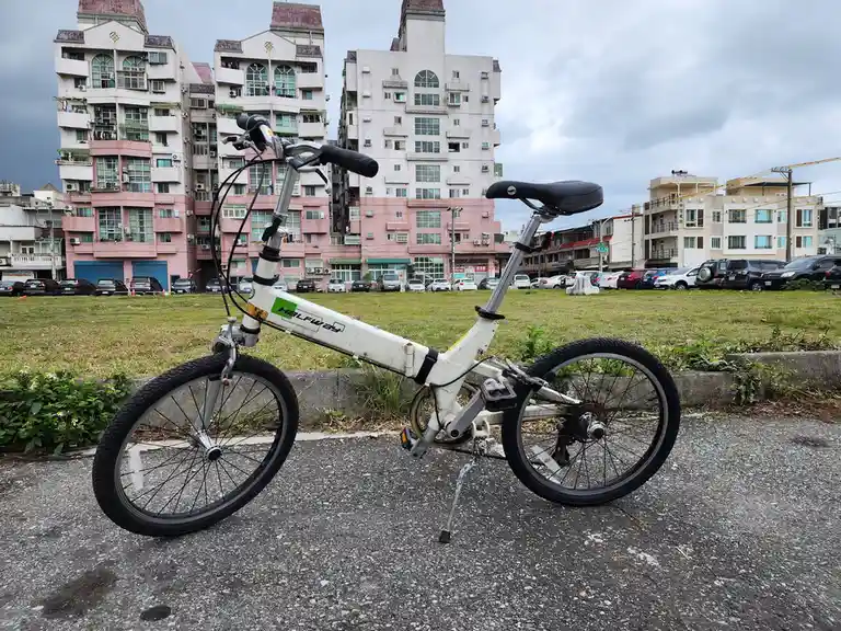 A white, foldable bicycle branded 'HALFWAY' is parked on a paved surface with its kickstand down, facing left. Behind it is…