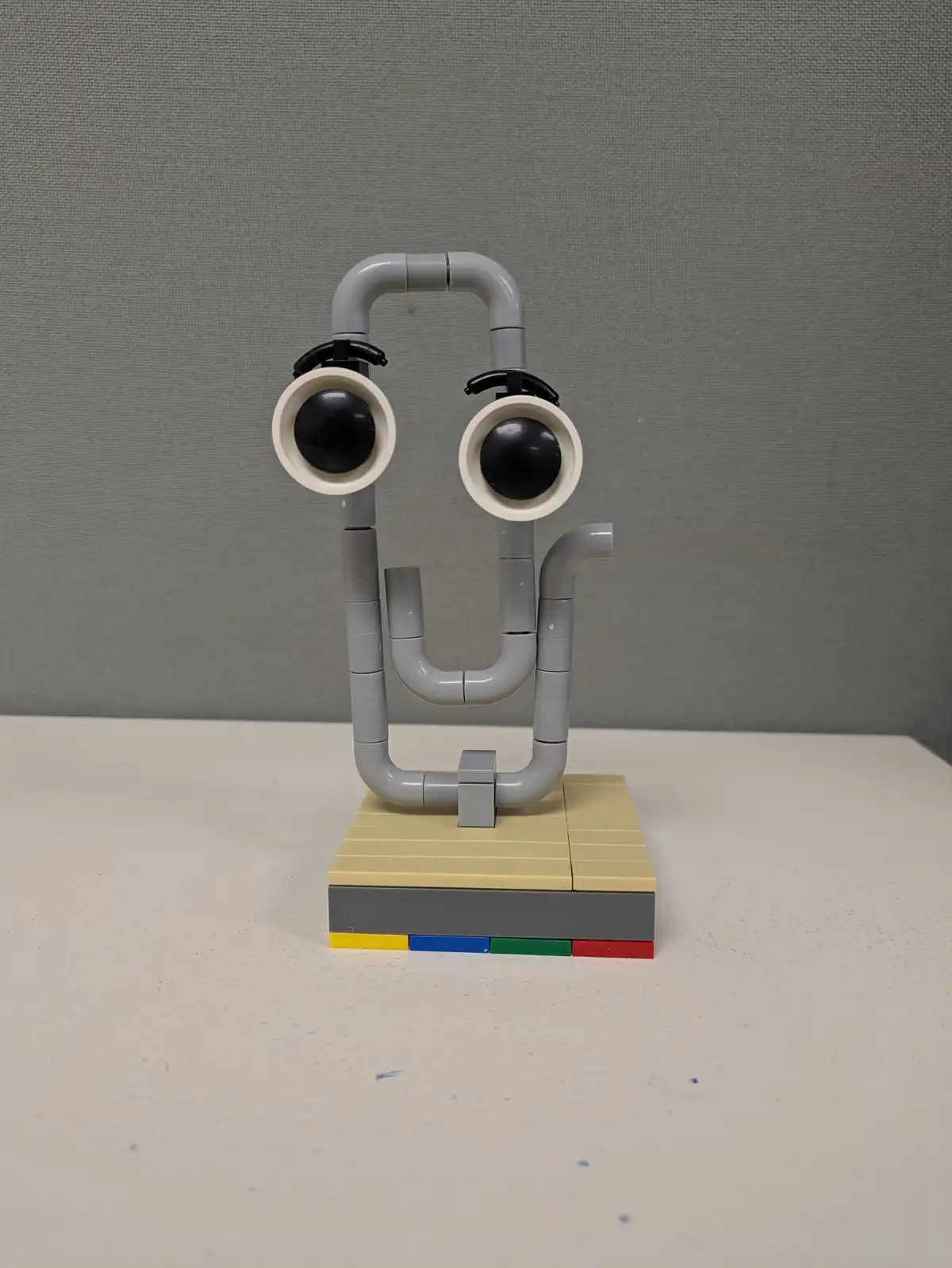 A Lego sculpture made of gray pipes and black-and-white googly eyes forms a face with a U-shaped mouth and a single pipe…