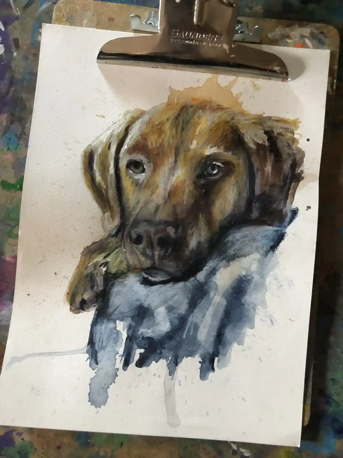 A watercolor painting of a dog's head on a white sheet of paper, clipped to a Saunders clipboard, shows the dog's face…