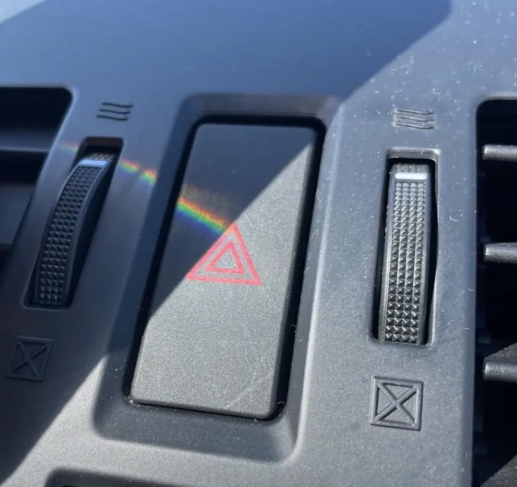 A close-up of a car's dashboard shows a hazard light switch with a red triangle symbol, where sunlight creates a rainbow…