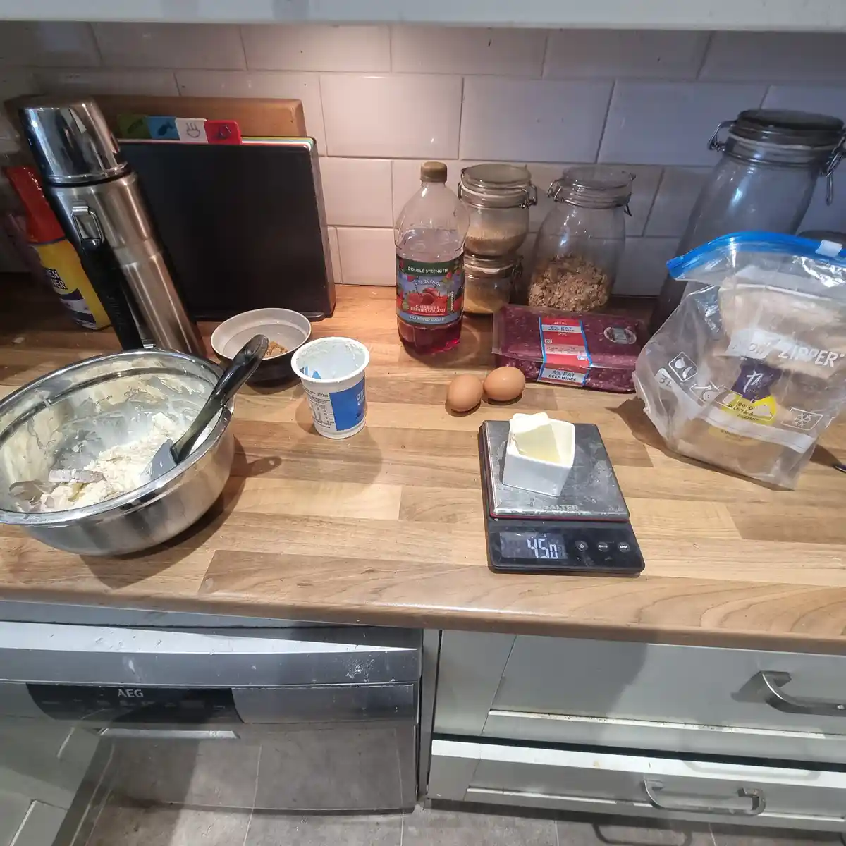 A kitchen counter displays baking ingredients including a mixing bowl, eggs, butter on a digital scale reading 450, flour…