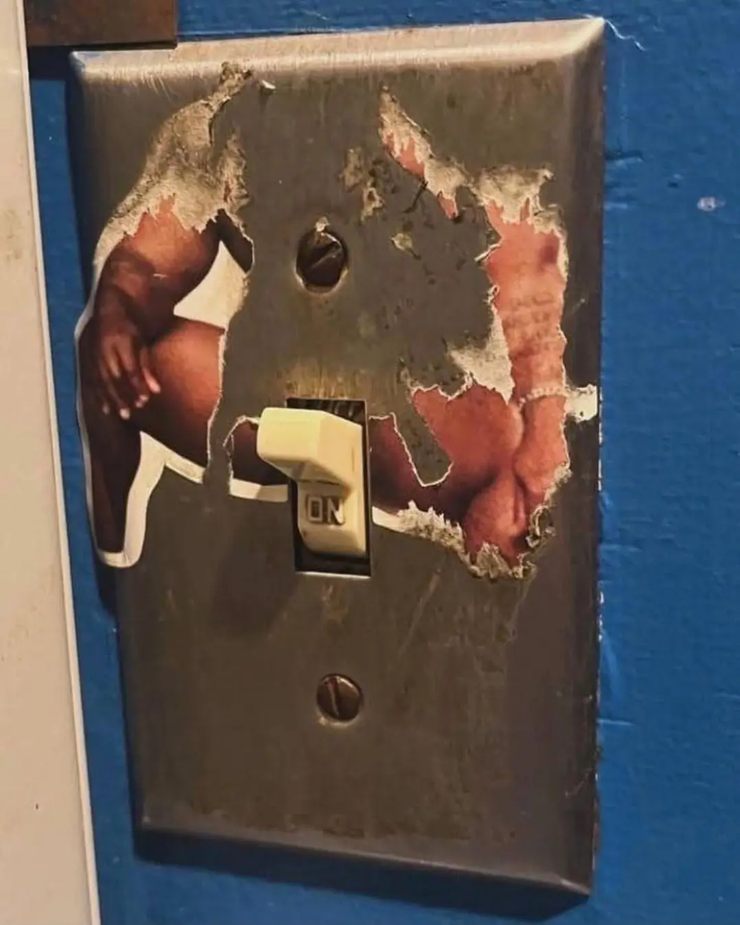 A weathered light switch plate with torn sections reveals a photo of a muscular man in white underwear underneath. The…