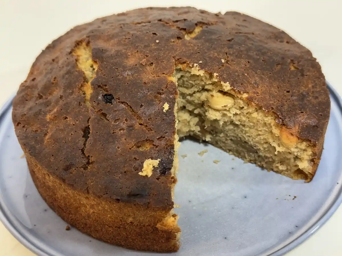 A round, slightly burnt-looking cake with a slice removed sits on a speckled blue plate. The cake's crumb appears dense and…