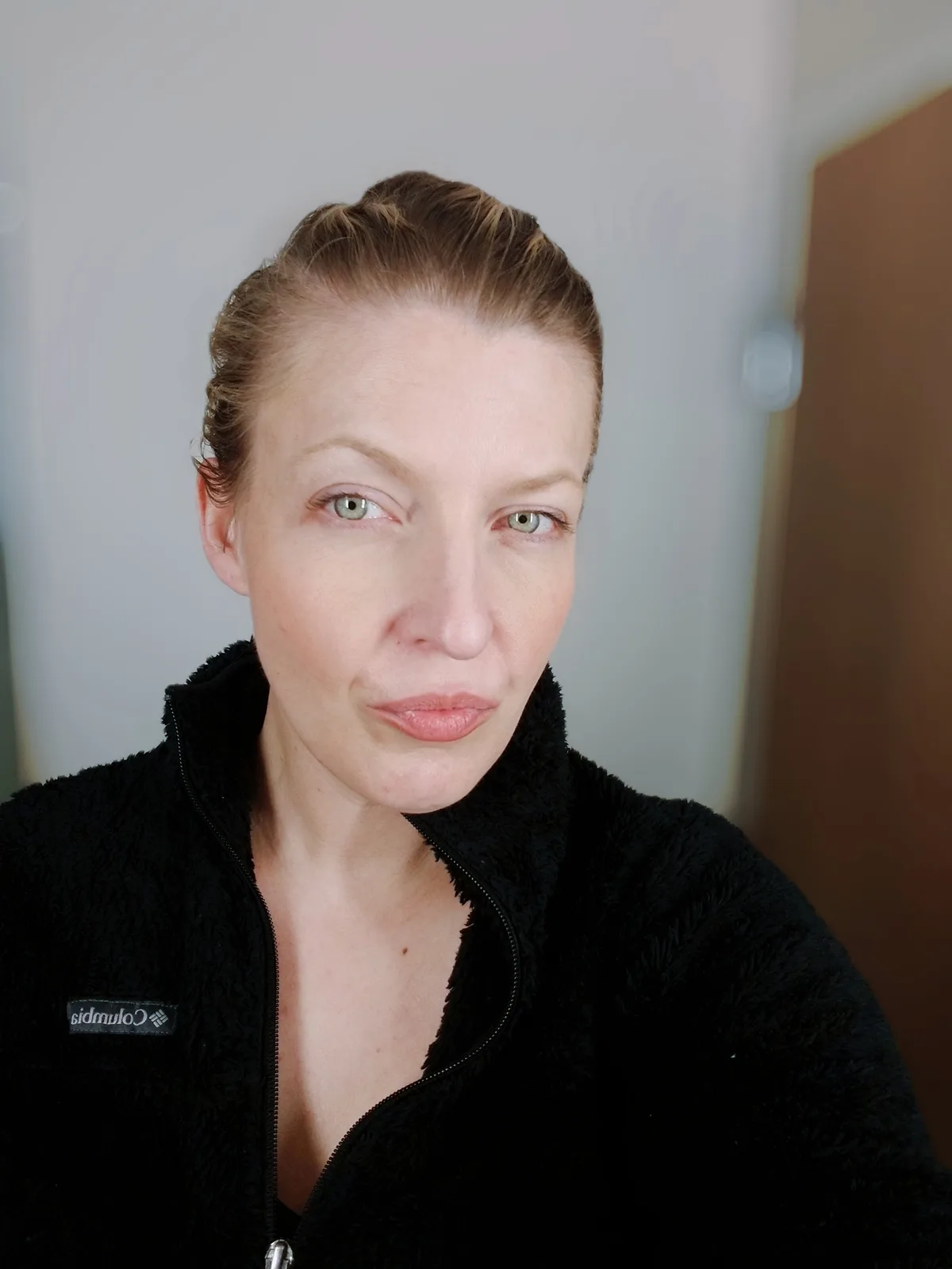 A woman with blonde hair pulled back and green eyes stares directly at the camera with a neutral expression while wearing a…