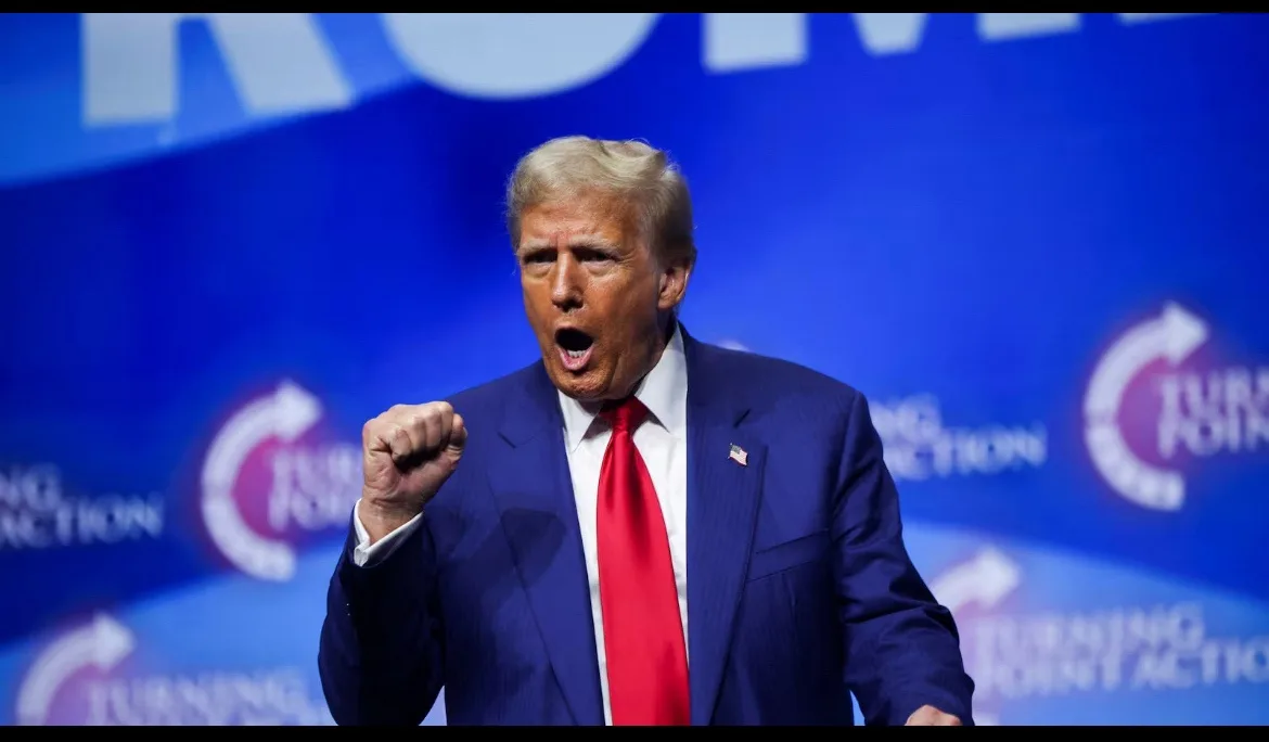 a man in a blue suit and red tie stands before a blue backdrop with 'turning point action' logos, his mouth open and right…