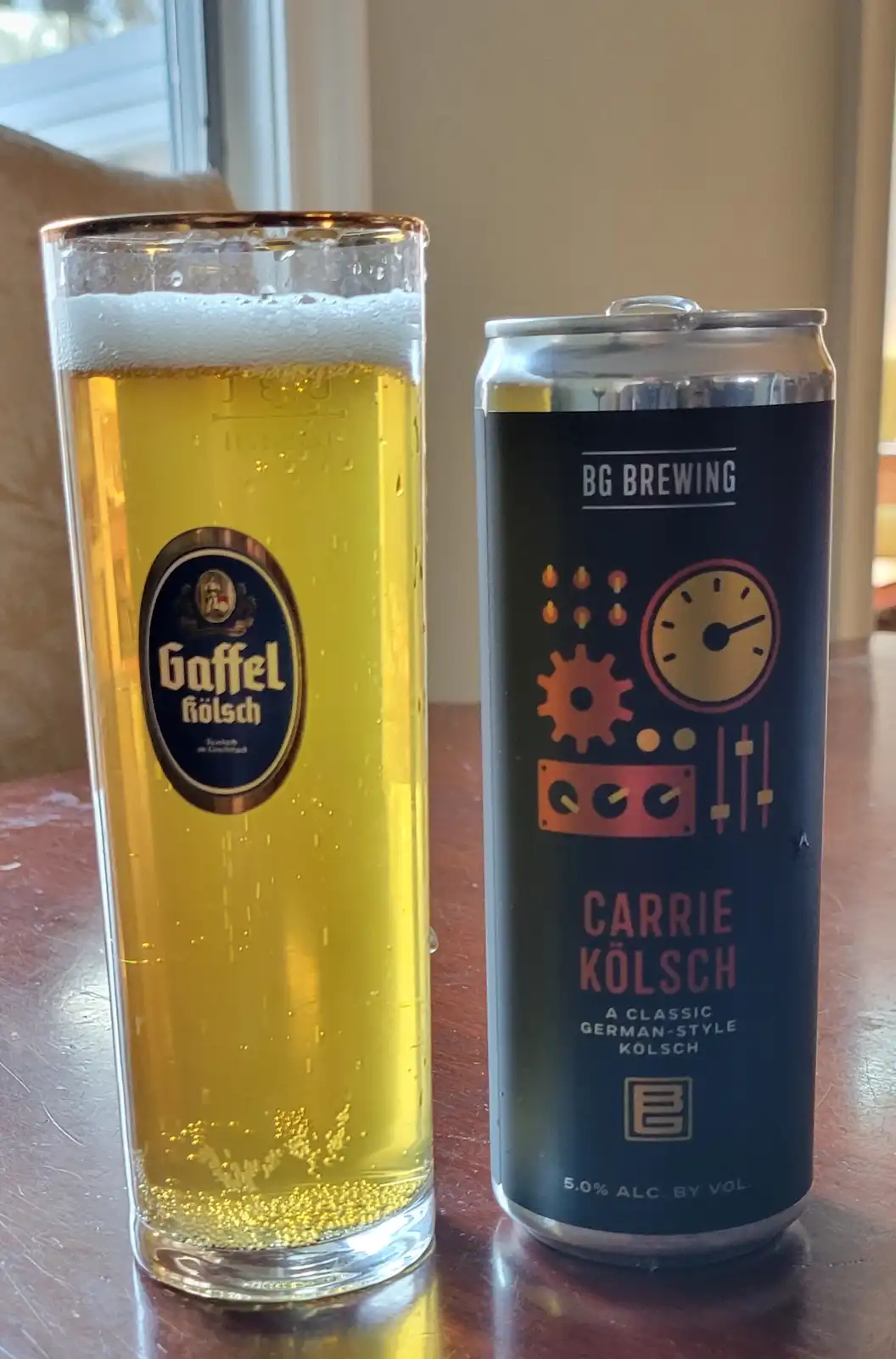 A tall glass of golden beer with a white head sits next to a dark blue can labeled 'BG BREWING' and 'CARRIE KÖLSCH'. The can…