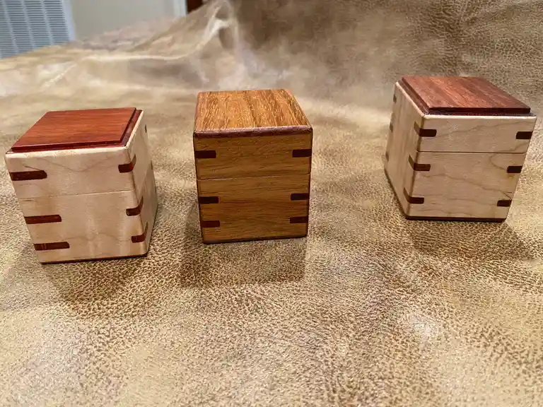 Three handcrafted wooden puzzle boxes of varying designs sit side-by-side on a textured beige fabric surface. Each box…