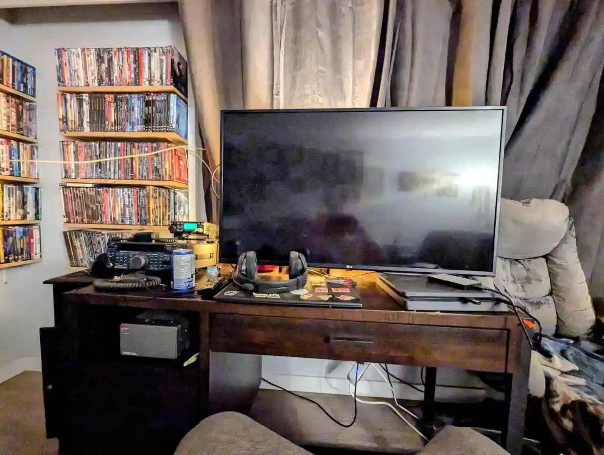 A room with a wooden desk holding a large TV, headphones, a laptop with stickers, and a gaming console. Behind the desk…