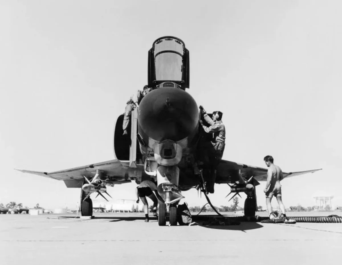 A black-and-white photo shows several men working on a fighter jet on an airfield. Two men are climbing on the aircraft's…