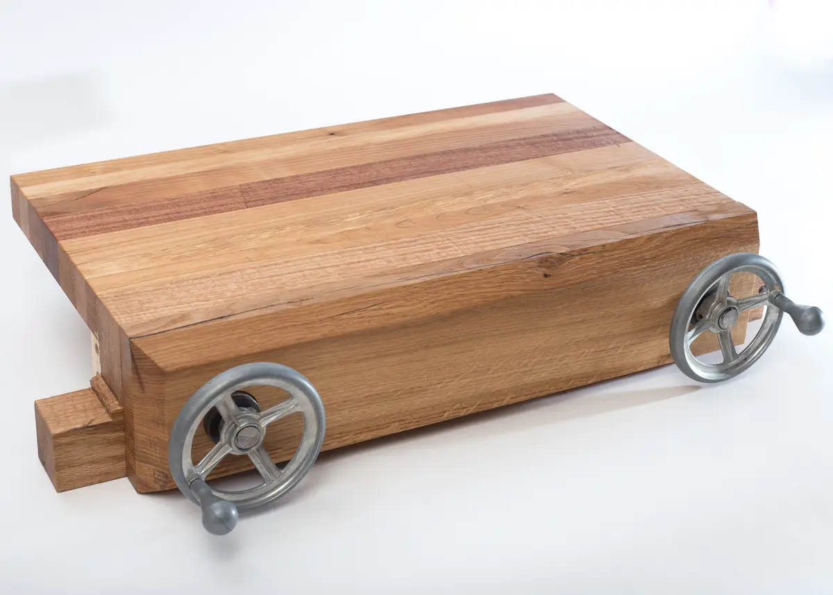 A wooden cutting board with two metal hand-crank wheels attached to its sides, resembling a tiny car. The overlaid text…