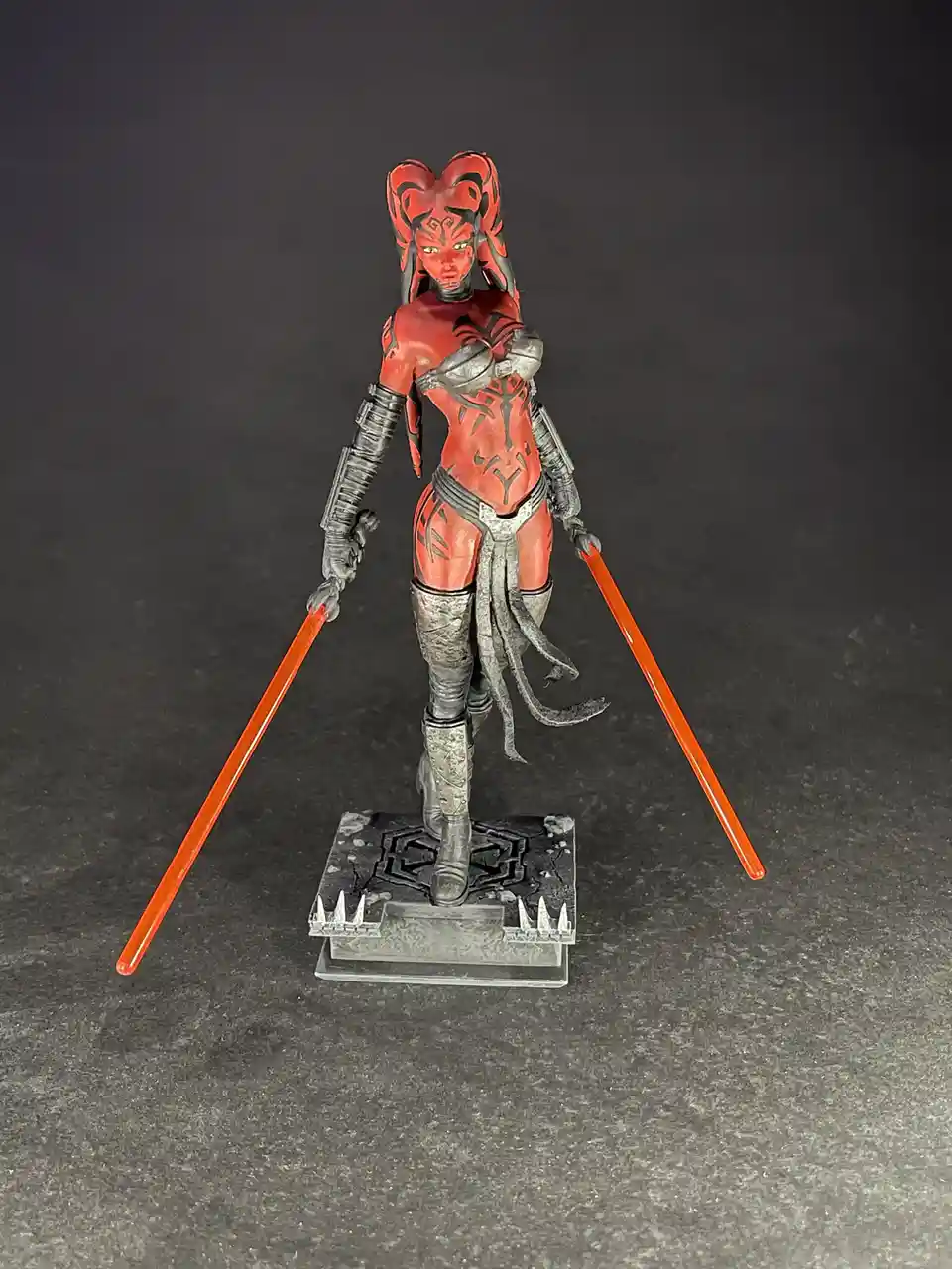 A painted action figure of the Star Wars character Asajj Ventress stands on a detailed base, holding two orange lightsabers…