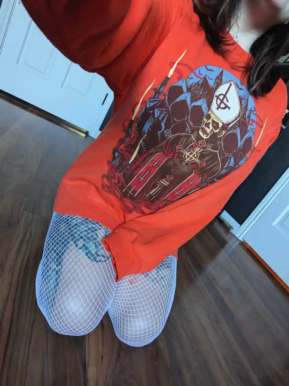 A person wearing a bright orange sweatshirt with a graphic of a skull wearing a bishop's mitre and holding a weapon, paired…