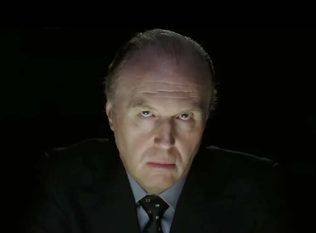 a close-up of a man in a dark suit and tie, staring intensely with narrowed eyes against a black background. The overlaid…