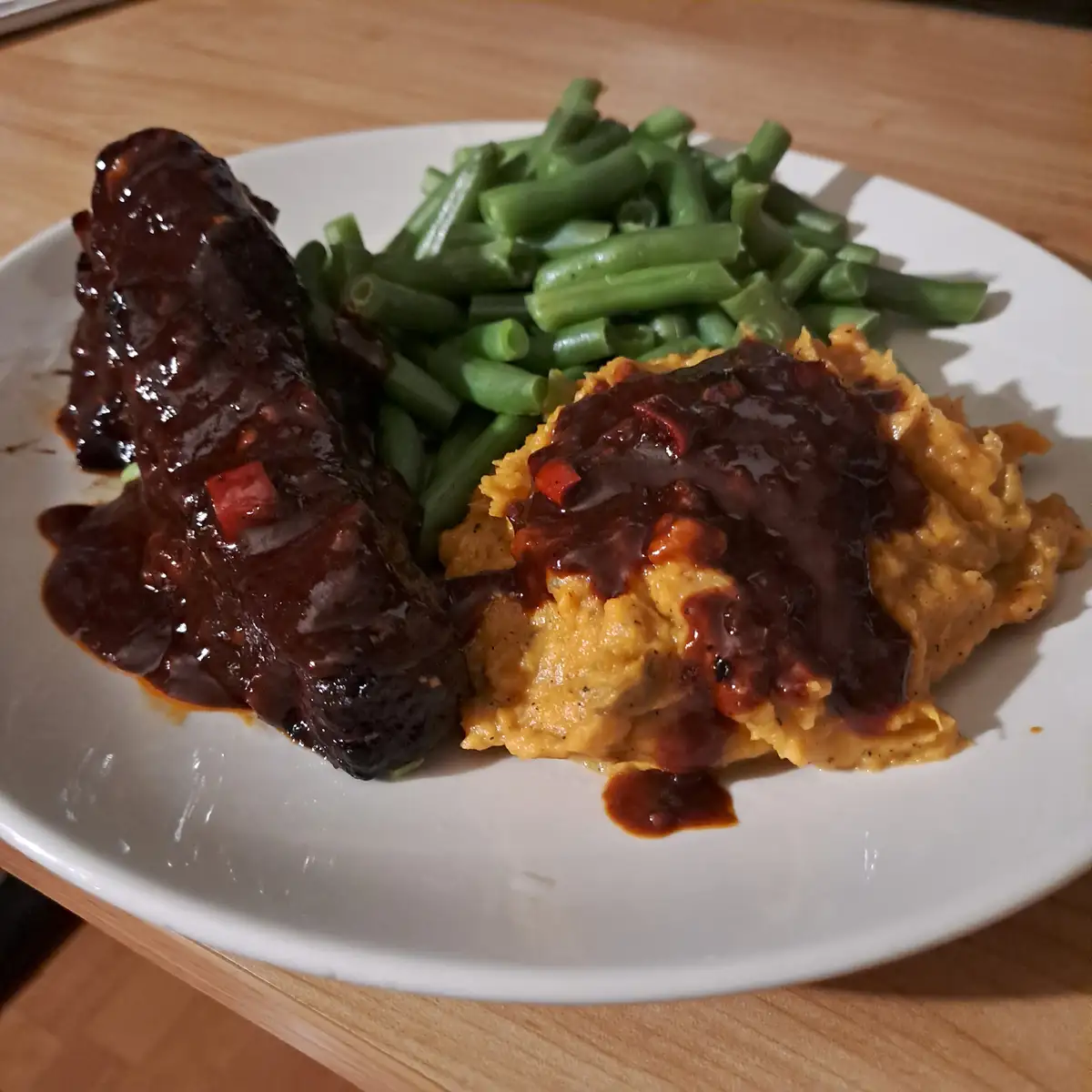 A close-up photo of a white plate on a wooden table holding a serving of ribs covered in dark sauce, a scoop of orange…