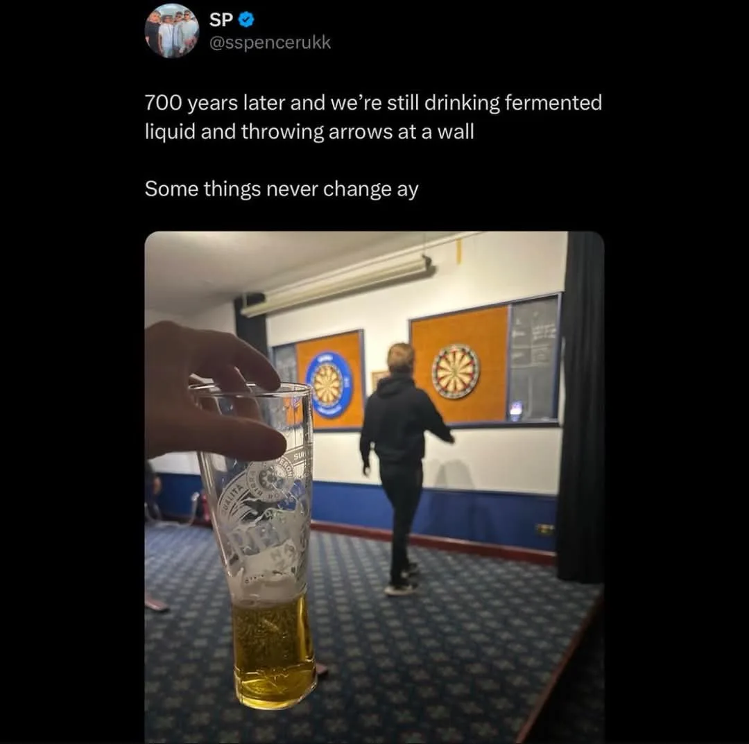 A hand holds a pint glass of beer in the foreground, while in the background a person in a black hoodie throws a dart at a…