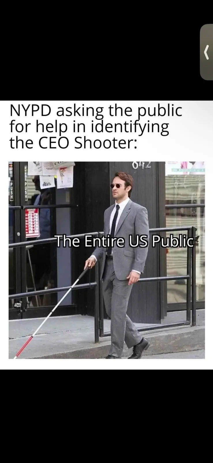 A man in a gray suit, white shirt, and black tie walks down a sidewalk while holding a white cane with a red tip. He wears…