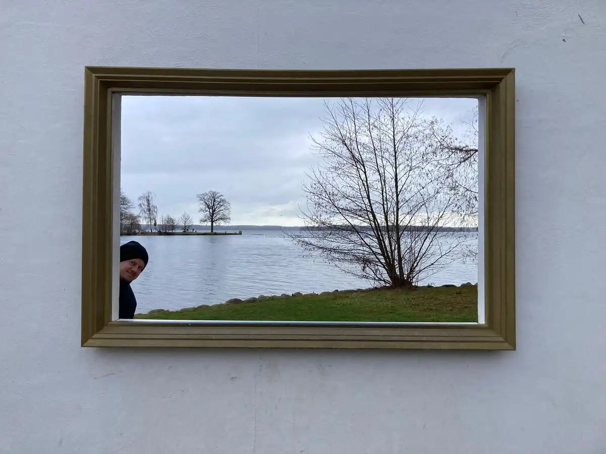 A person wearing a black beanie peeks out from the left side of a gold picture frame mounted on a white wall, while the…