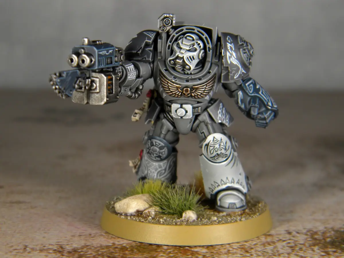 A heavily armored, silver-and-gray painted miniature of a Space Marine from Warhammer 40,000 stands on a circular base with…