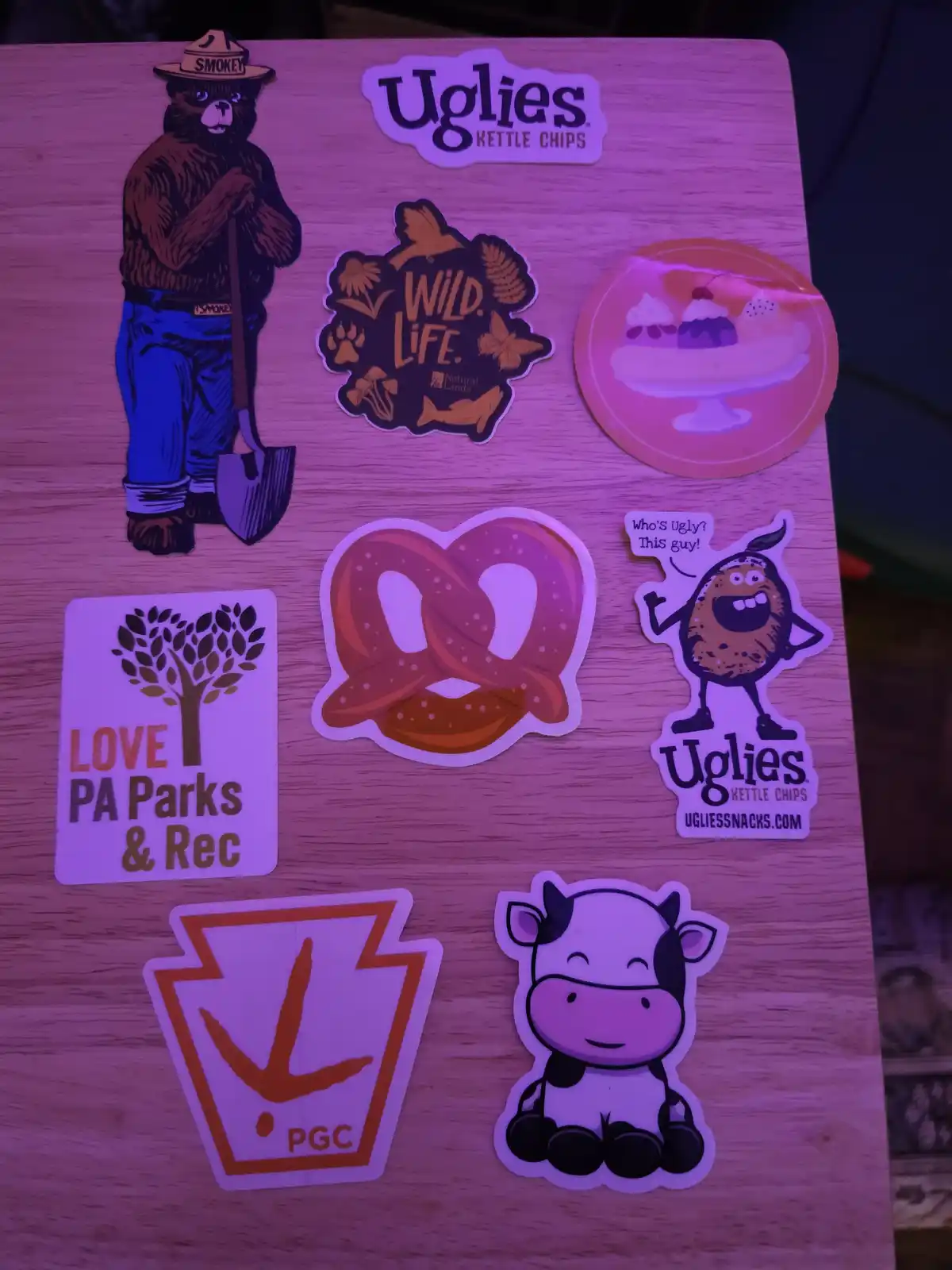 A collection of stickers on a wooden surface under purple lighting, including Smokey Bear, 'Uglies Kettle Chips' branding, a…