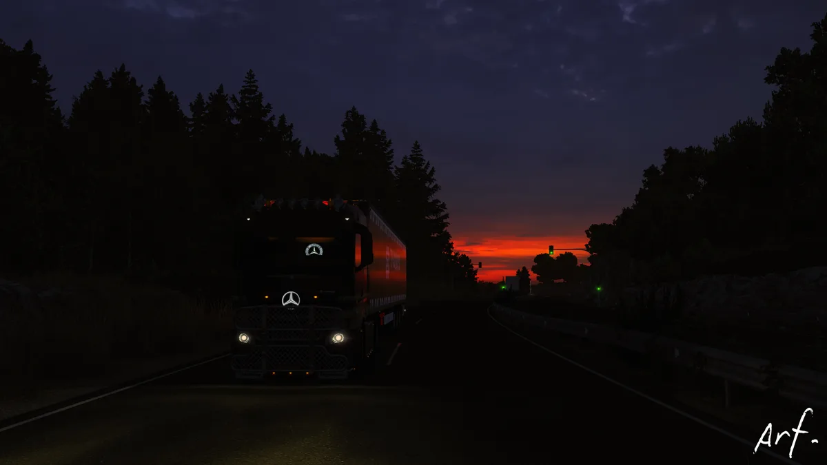 A dark Mercedes-Benz truck with headlights on drives toward the viewer on a road at dusk, with a vibrant red-orange sunset…