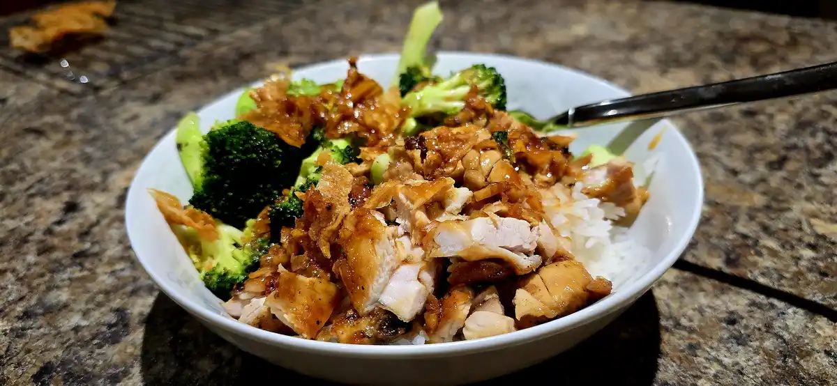 A close-up photo of a white bowl filled with rice, glazed shredded chicken, and steamed broccoli, resting on a granite…