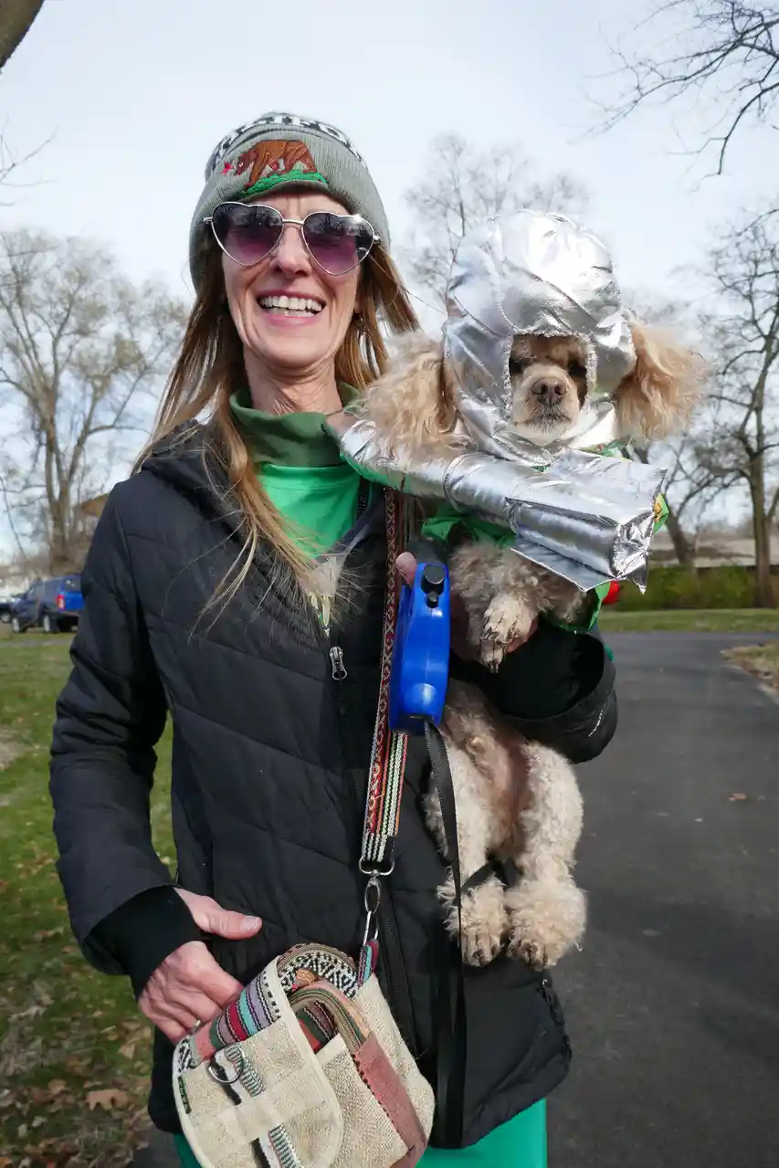 A woman wearing a grey beanie with a bear logo, heart-shaped sunglasses, and a black puffer jacket smiles while holding a…