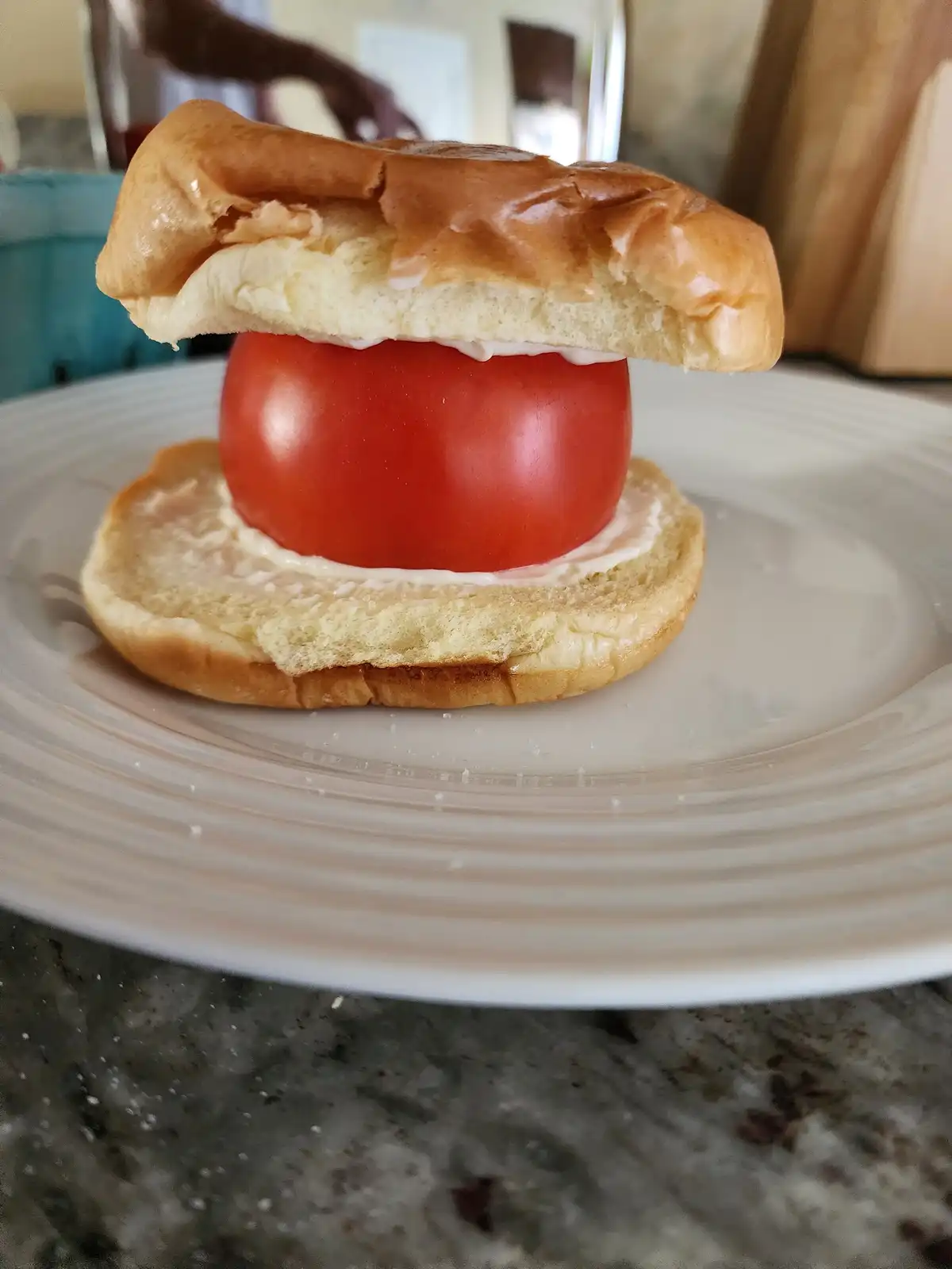 A close-up photo shows a sandwich made with a single large red tomato between two halves of a soft, golden-brown bun, both…
