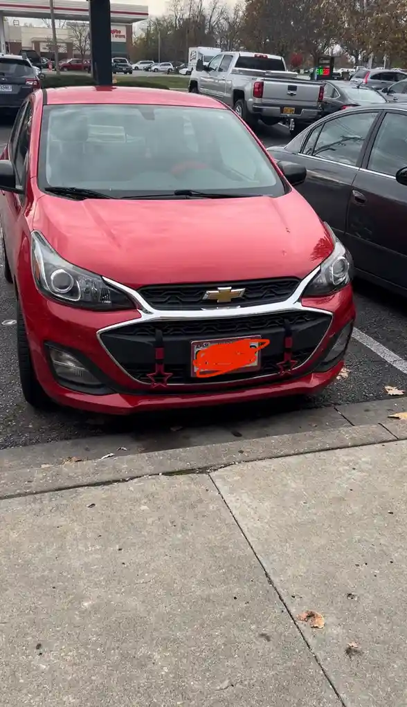 A red Chevrolet Sonic is parked in a lot with its license plate obscured by orange scribbles. The car has a black grille…