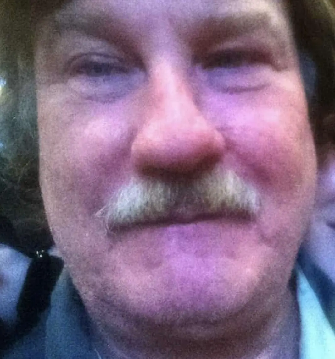 A close-up, low-resolution photo of a man with a mustache and headphones, looking slightly squinted or unimpressed. The…