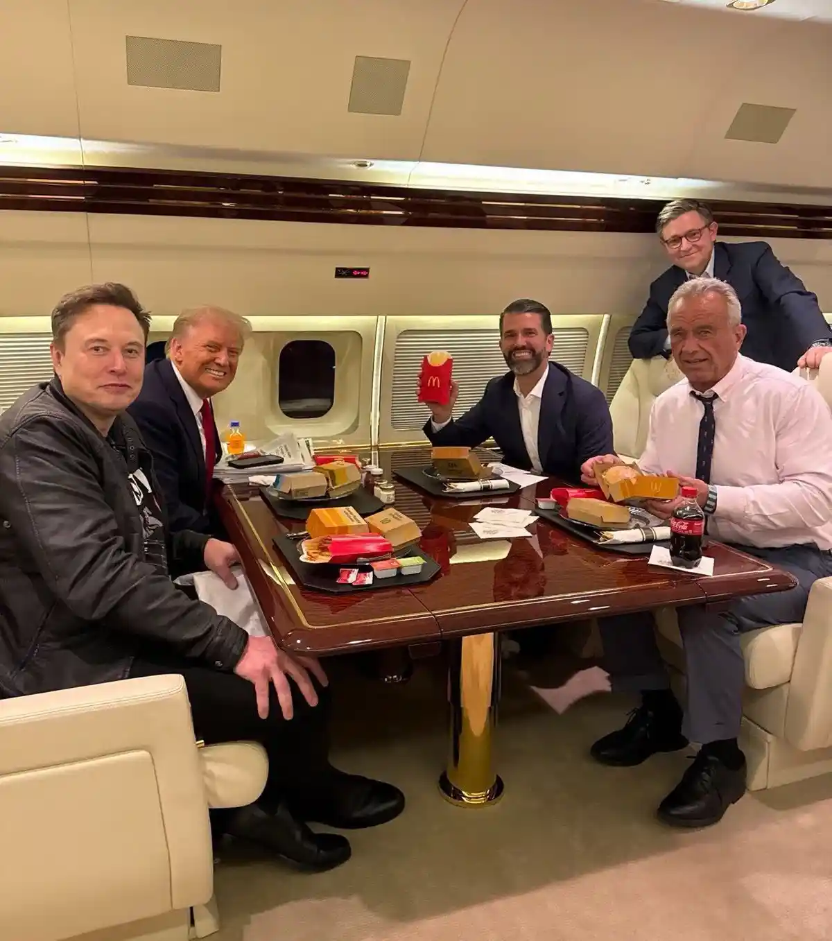 Four men are seated around a polished wooden table inside a luxurious private jet cabin, eating McDonald's meals. Elon Musk…